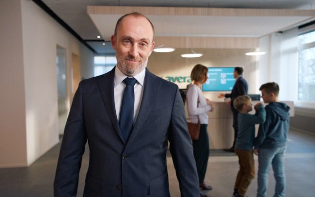 The main actor if the commercial for the bank avera commercial standing in a branch of the bank in front of people.