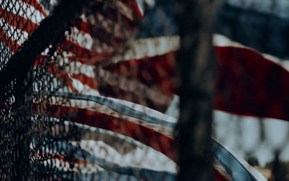 A lot of american flags wagging in the wind behind a fence at a rodeo competition on a sunny day.