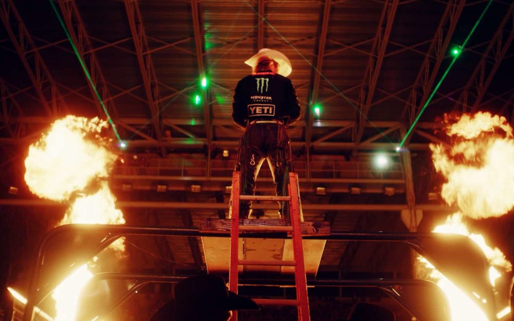 The bullrider Derek Kolbaba standing on a podium between fire and lights before a competition.