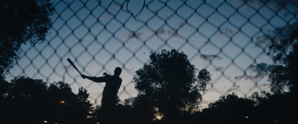 A shiluette of a boy with a baseball bat hitting a ball behind a fence in the evening.