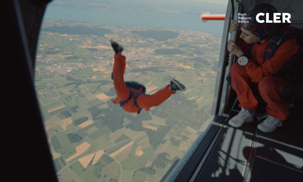 A person in a orange jumpsuit doing a skydive out of a plane while another person watches.