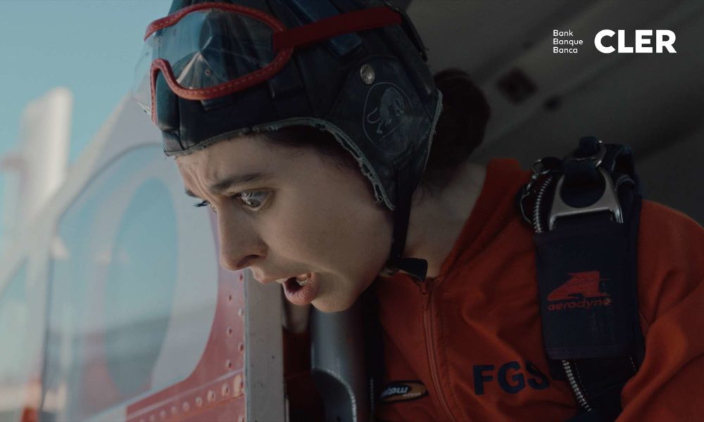 A woman in an orange jumpsuit whith a helmet looking out of a flying airplane.