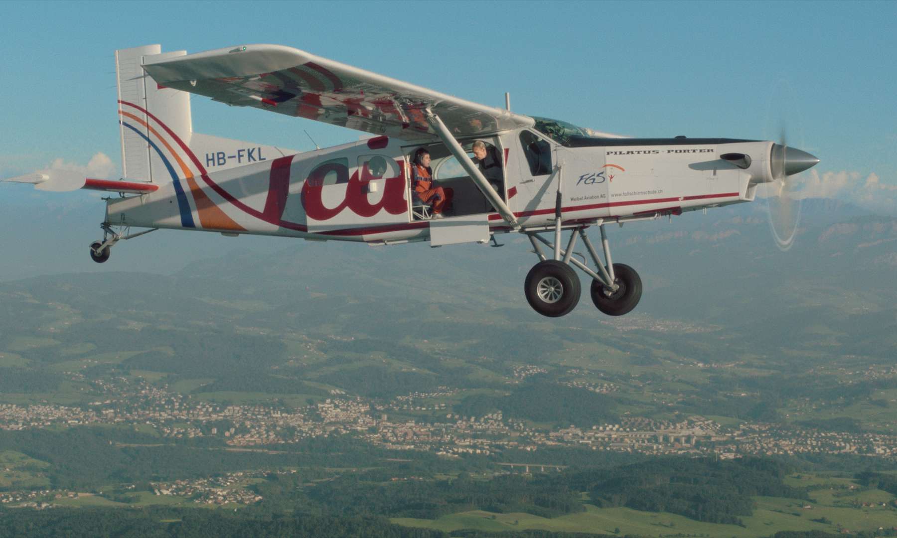An airplane with some skydivers in it and an open door flying over zurich countryside.