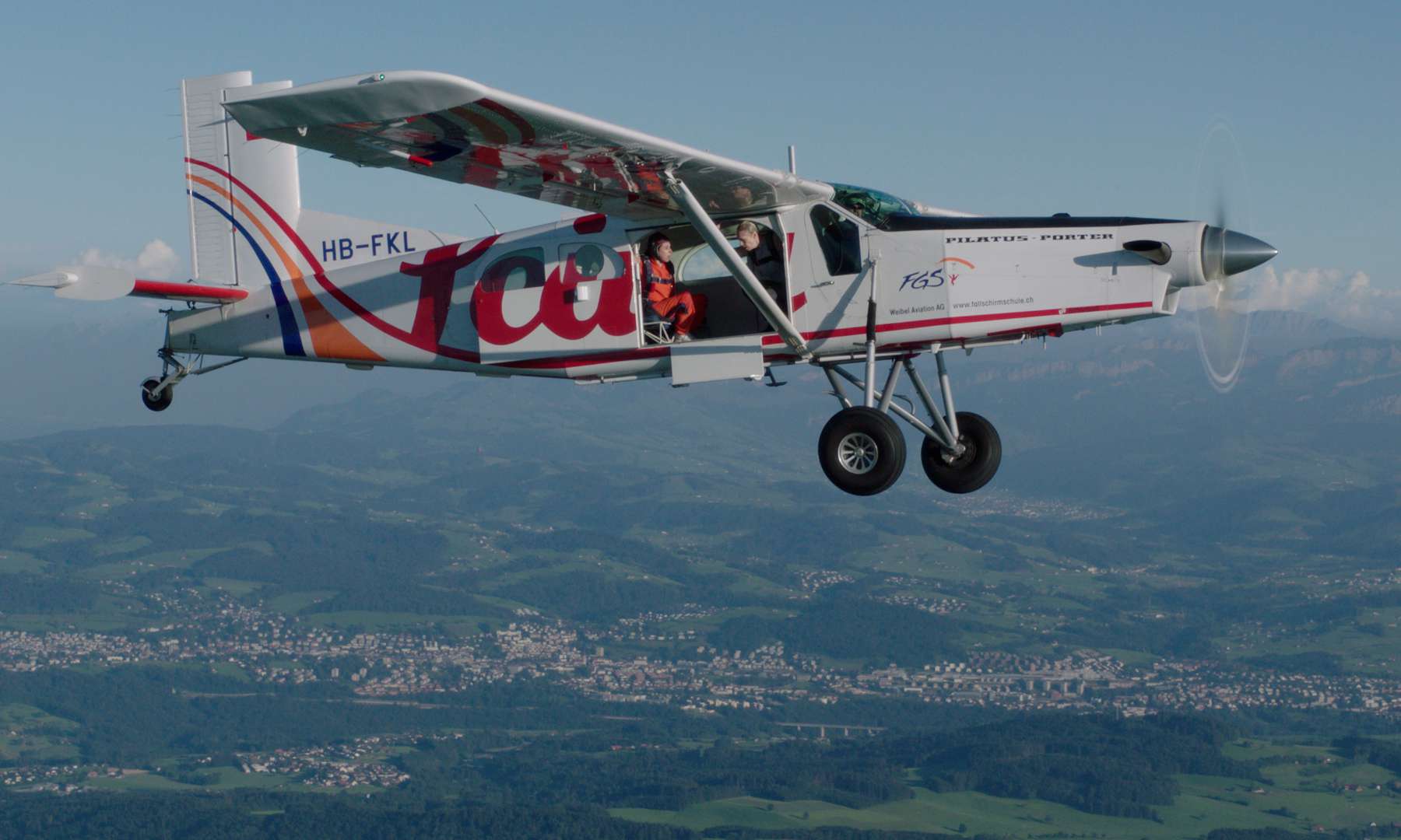 An airplane with some skydivers in it and an open door flying over zurich countryside.