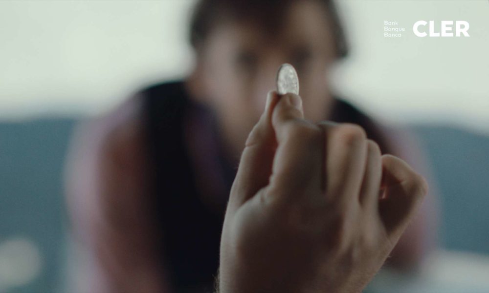 A hand holding one swiss franc up, while another person in the background is watching.