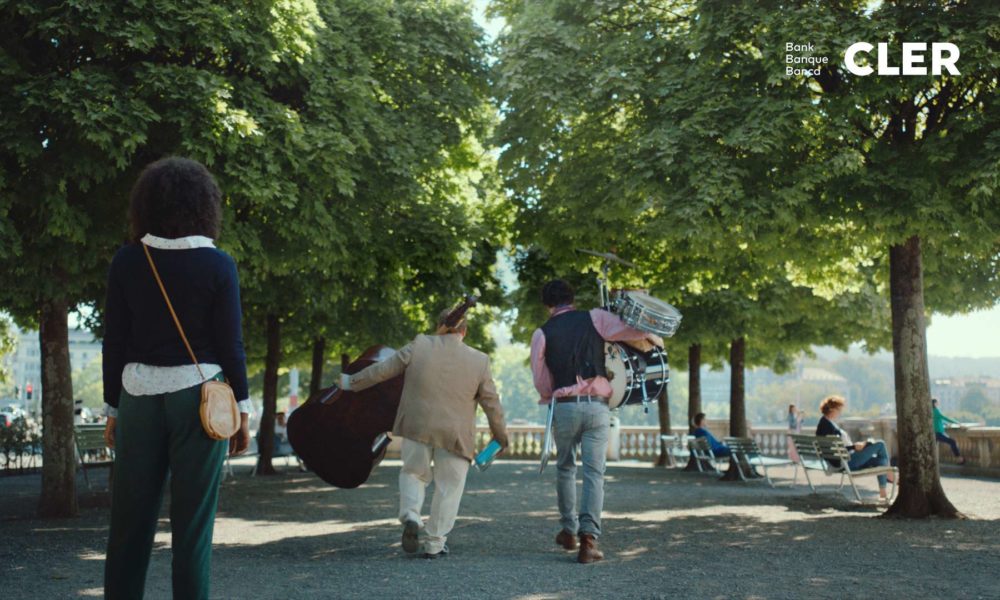 Two musicians carrying instruments walking beneath trees while a woman is watching them.