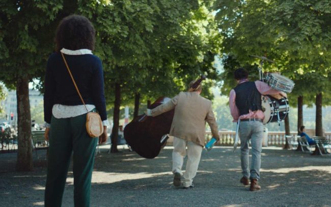 Two musicians carrying instruments walking beneath trees while a woman is watching them.