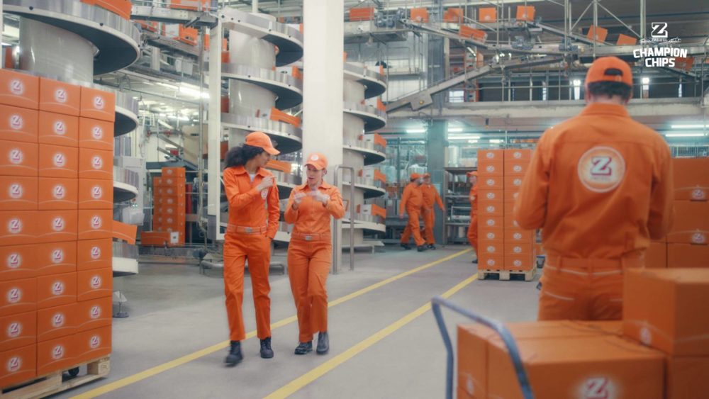 Two persons walking through a massive produciton hall in orange suits.
